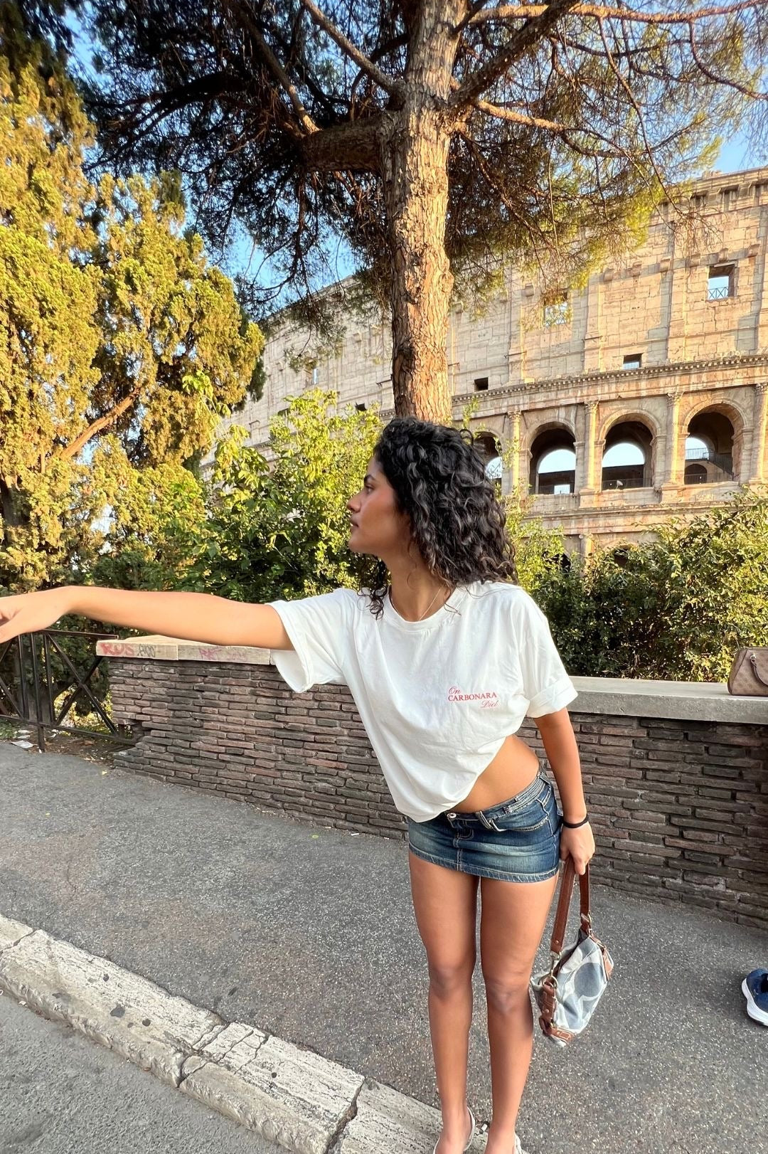 Woman standing in front of the Colosseum with trees and a clear sky.